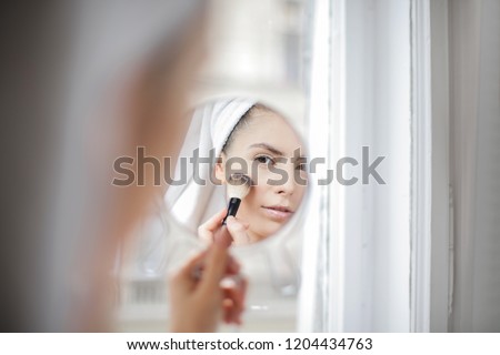 Young woman looking herself in the mirror and applying power on her face.