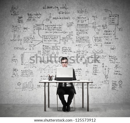 Businessman Working At A Laptop With In The Background A Wall Full Of Economy Drawings