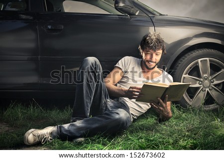 handsome boy reading a book lying on the grass with beautiful car behind him