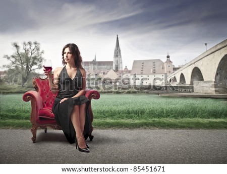 Elegant woman sitting on an armchair and holding a glass of wine with antique village in the background