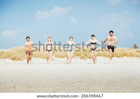 A summer day on a sandy beach, a group of five young people run towards the sea to swim. People are in swimwear on the beach and there are no other people.