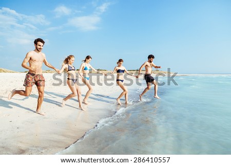 A summer day on a sandy beach, a group of five young people run towards the sea to swim. People are in swimwear on the beach and there are no other people