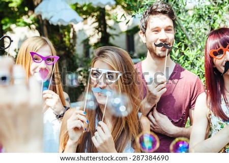 Party! A group of friends, three women and a man have fun at a party in a park with a mustache and fake glasses, joking and talking to each other and playing with soap bubbles in the air!