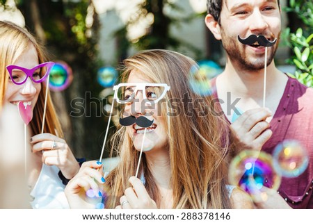 Party! A group of friends, two women and a man have fun at a party in a park with a mustache and fake glasses, joking and talking to each other and playing with soap bubbles in the air!