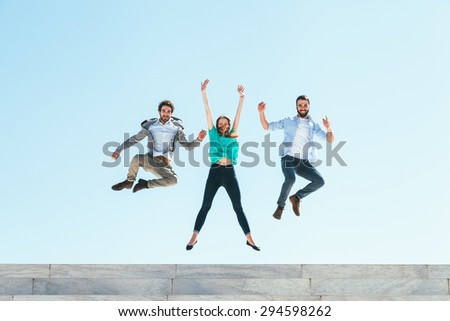Three friends, two men and a woman, jump on top of the steps of an amphitheater, behind them the azure sky of a sunny spring day
