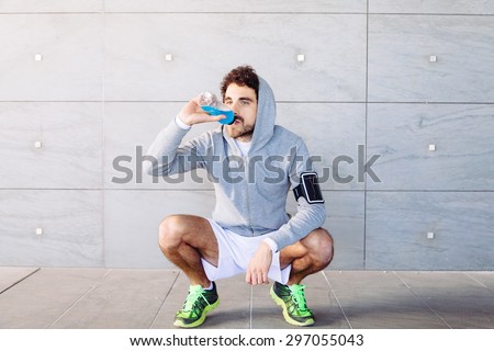 A young man in sportswear resting in the shade of a building in a summer day, drinking an energy drink blue color from a bottle before resuming training