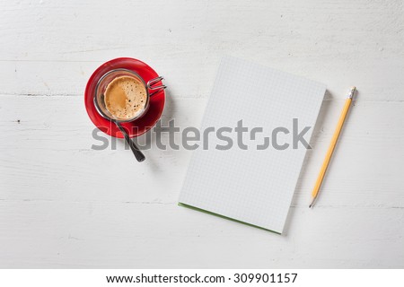 A recycled paper notebook checked with a black pencil with the eraser at the top and a cup of coffee with red saucer and spoon, are arranged on a wooden table painted white. View from the top