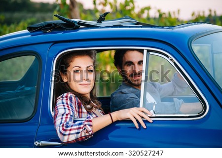 Summer end day, during sunset, a young couple in love lies on a blue vintage car during a trip to Tuscany, among rows of vineyards and olive trees