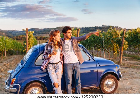 A loving couple watch the sunset, standing leaning against an old blue car in Tuscany, Italy. Around them, rows of vines and the classic hills. The young man shows to his girlfriend sunset