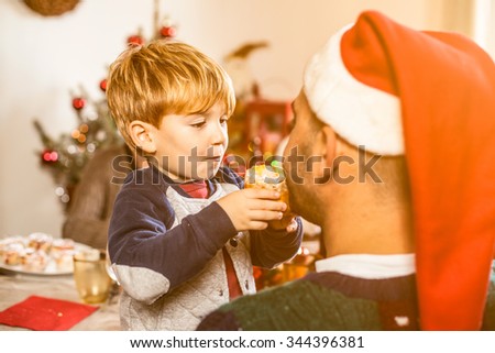 Father and son on Christmas Day. The child approaches the mouth of the father a small panettone to make it taste. The father has a red Christmas hat. Behind them on the table and the Christmas tree