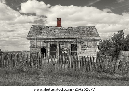 horizontal black and white  image of an old abandoned little house with a broken wooden fence in front of the house on a warm autumn day.
