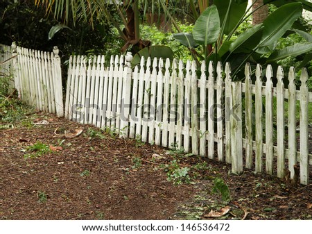 Moldy old white broken picket fence in front of a green garden.