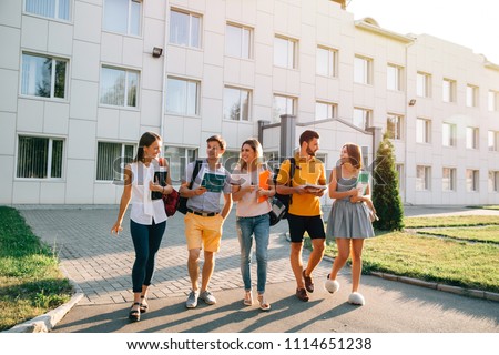 Free time of a students, bachelor`s campus life rhythm. Five friendly students are walking after they passed test outside the college building and discuss the project, smiling, enjoying, carefree