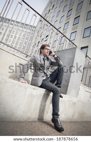 A young businessman sitting on the street and talking on the phone on the backdrop of skyscrapers 		 A young businessman in the street talking on the phone