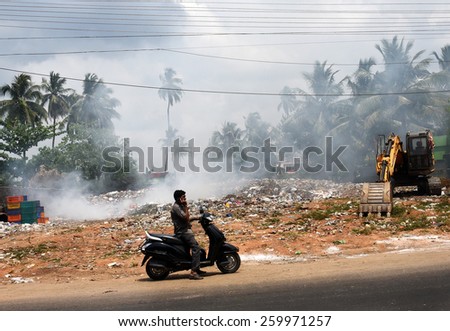 TRIVANDRUM, KERALA, INDIA, MARCH 04, 2015: Land and air pollution in India. Environmental hazard. Garbage being burnt in the open, a health hazard. A man on scooter stops to talk on his mobile phone.