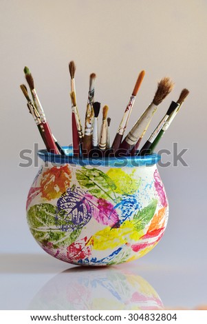 Art work on ceramic pot. Brightly colored Stencil images of leaves on pot painted white. Paint brush holder. Artist\'s workshop. Leaf impressions on earthen pot.