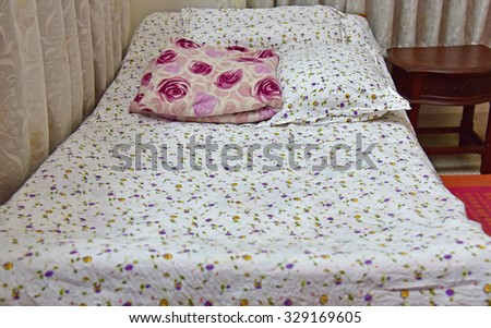 Bed covered with white printed bed-sheet. Pillows and a woolen blanket are also seen.