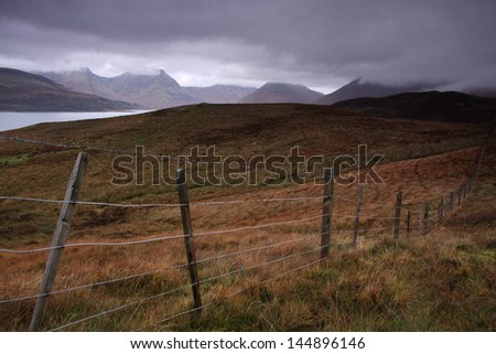 A wire fence leads into the distance through moorland by Loch Slapin. Looking towards Bla Bheinn and the Cuillin Hills. Low grey clouds obscure mountains. Isle of Skye, Scotland. Midday in October.