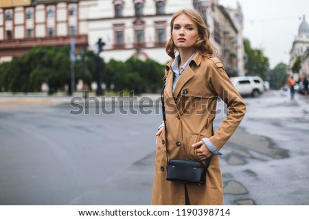 Young attractive woman in trench coat with little black cross bag dreamily looking in camera spending time on cozy city street