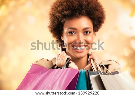 Stylish African-American woman posing in front of a glittering gold background ang holding shopping bags.