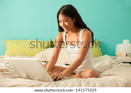 Young Asian woman sitting on her bed and chatting with someone on the Internet on her laptop.