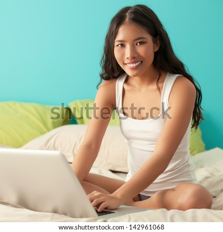 Young Asian woman sitting on her bed and browsing through the Internet on her laptop.
