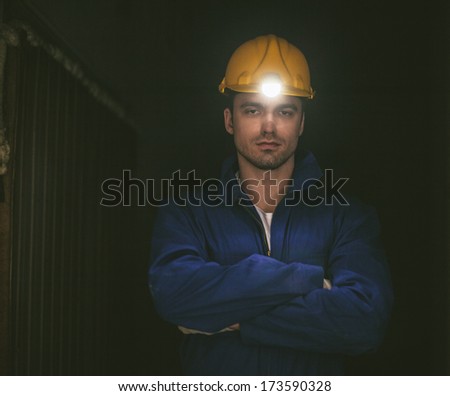 Dark indoor portrait of a young builder wearing a yellow hardhat.
