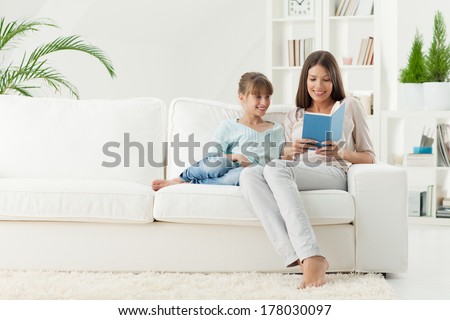 Smiling mother and daughter reading a book together in the living room.
