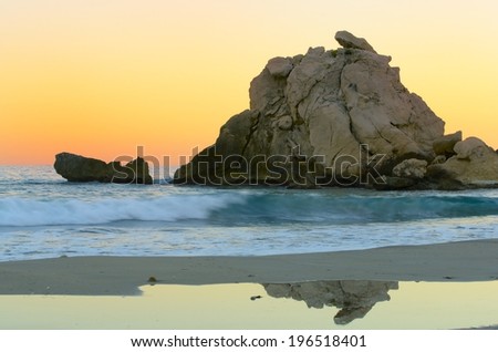 beach with turquoise sea, orange sky and reflections in the sand, with a large rock in the sea. Idyllic beach.