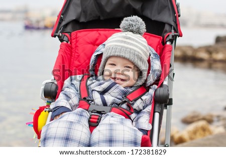 happy baby boy sitting in a red stroller for a walk in the background of the sea
