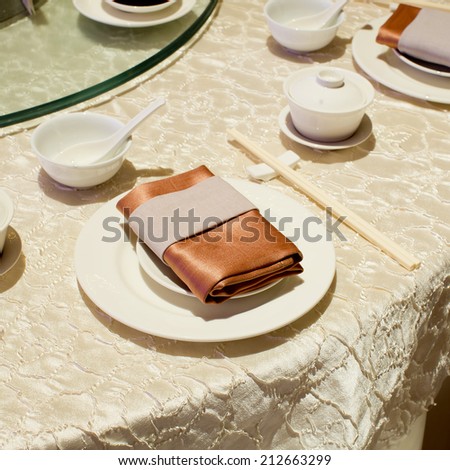 Brown napkin on blank plates, empty cup, soup bowl, spoon and chop stick on dinner table