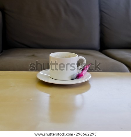 cup of coffee with sugar pack and coffee spoon on wood table with black sofa background.