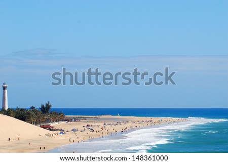 A sunny sand beach being hit gently by foamy wave crests, with a lighthouse standing on a pouch of tropical trees, with a vast Atlantic Ocean in the horizon, in Fuerteventura, Canary Islands.