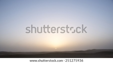 The  sun is about to fall behind the blue horizon in the Sahara desert in Morocco, with dunes in the front.