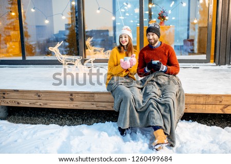 Young couple warming up with plaid and hot drinks sitting on the terrace of the modern house in the mountains durnig the winter holidays