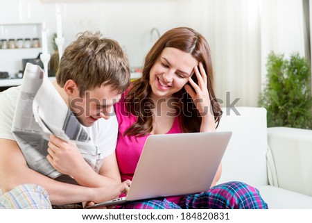 Young married couple in pajamas sitting in the sofa reading newspaper and laughing at home in the morning