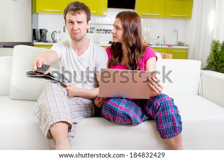 Young married couple in pajamas sitting in the sofa with newspaper and laptop and watching TV at home