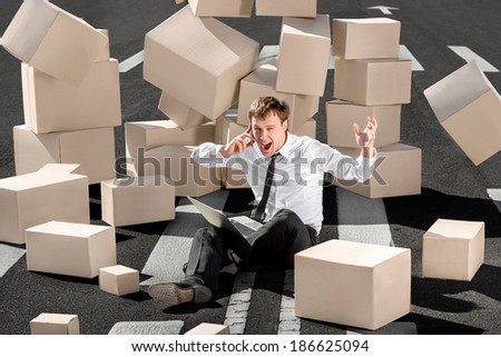 Crying and shocked businessman speaking phone and sitting with laptop on the asphalt road surrounded by falling carton boxes