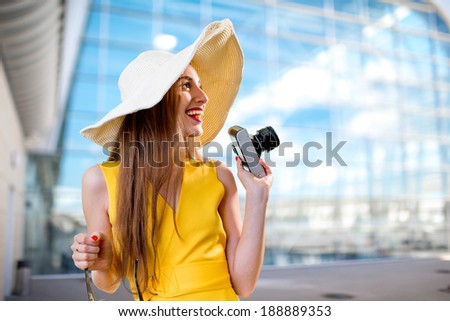 Young traveling woman with photo camera and panama dressed in yellow dress standing in front of the airport or high-tech background and looking toward