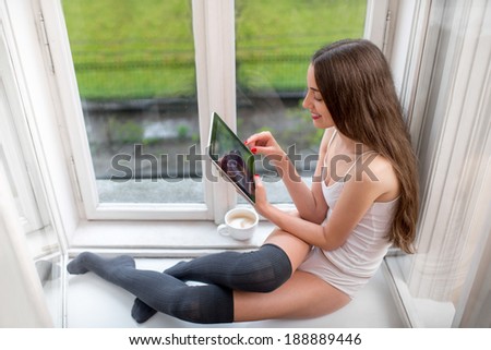 Smiling young woman reading something on the tablet and sitting on the windowsill at home