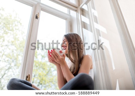 Young and cute lady sitting on the windowsill and looking out the window with cup of coffee in the morning