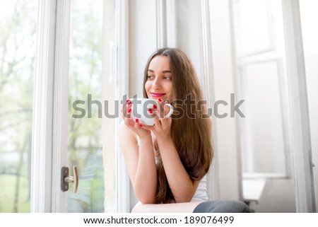 Young and cute lady drinking coffee on the windowsill and looking out the window