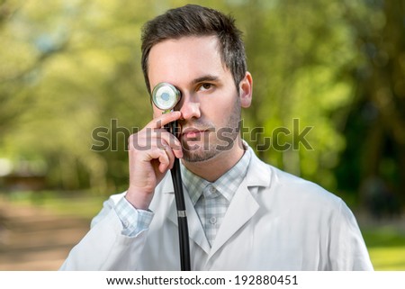 Crazy doctor with stethoscope instead his eye on green background in the park