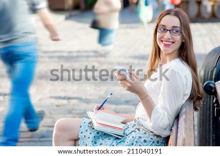 Young woman sitting and studying on the bench with books typing message on the phone in the city