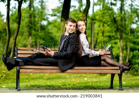 Young Business couple sitting on the bench and reading or working with tablets outdoors.
