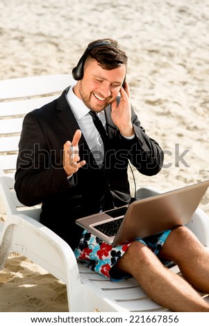 Businessman dressed in suit and shorts having video call with laptop on the sunbed at the beach