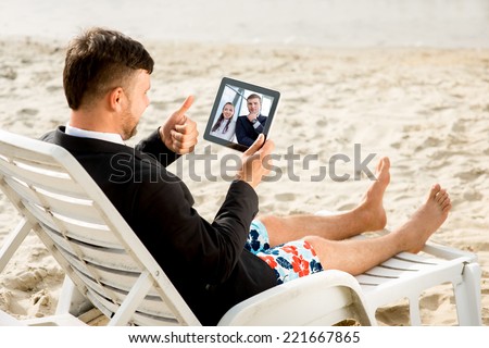 Businessman dressed in suit and shorts having video call with digital tablet on the sunbed at the beach