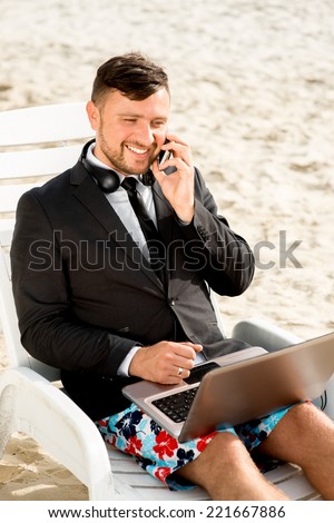 Businessman dressed in suit and shorts working with laptop on the sunbed at the beach