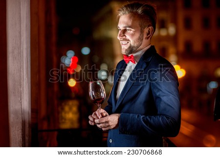 Smiling well-dressed man with wine glass standing on the balcony on the night city background