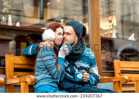 Young loving couple dressed in blue sweater sitting with knitted coffee cups on the bench near cafe in winter